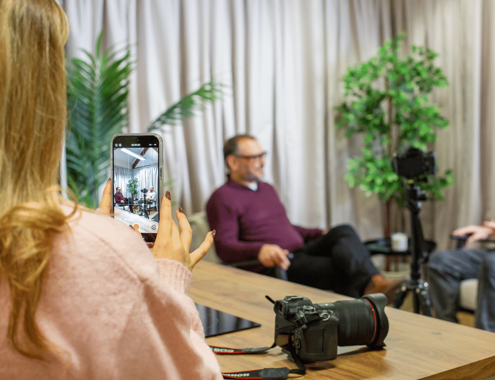 A woman filming a man on her phone, with a camera on the table in front of her