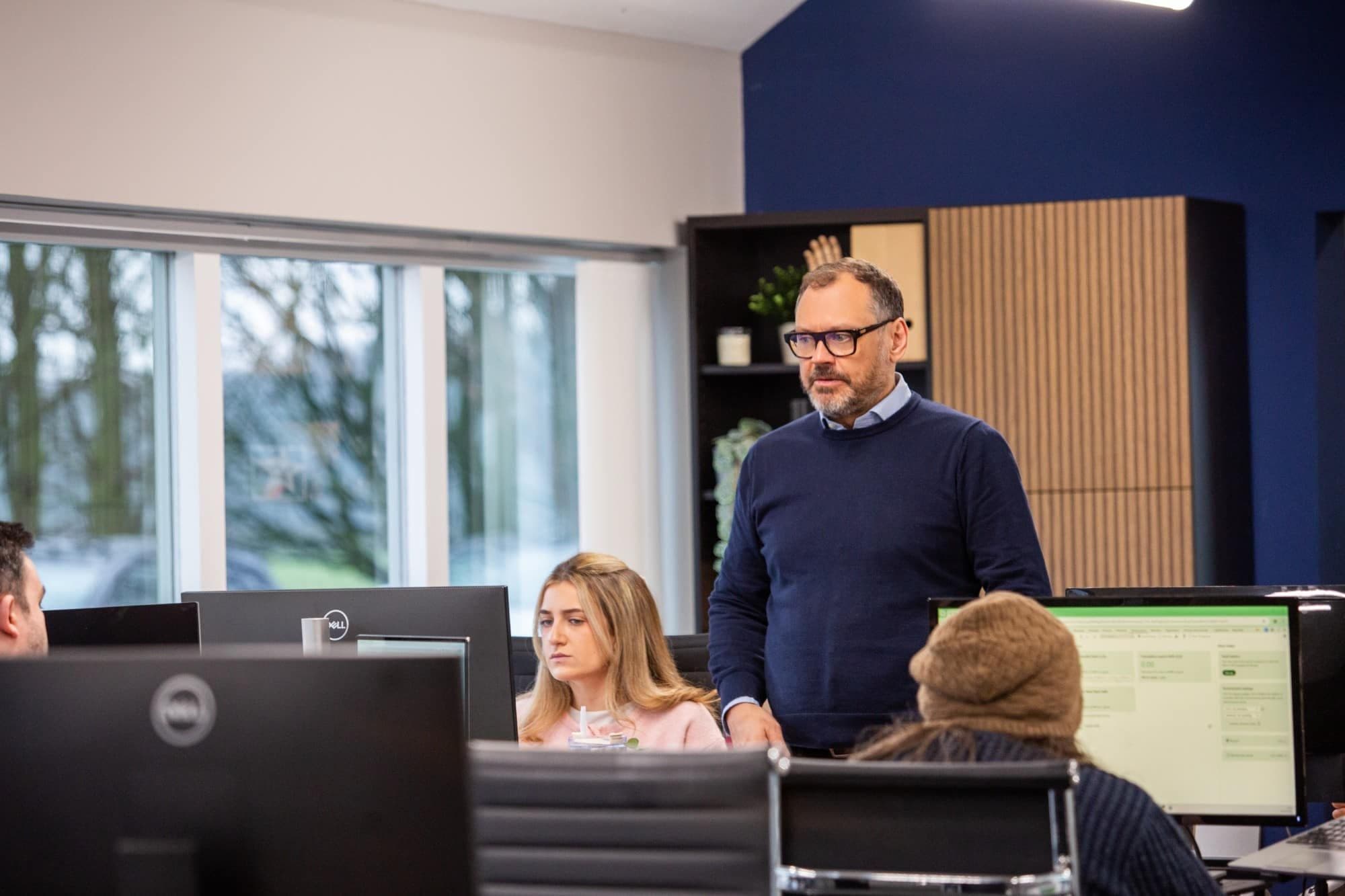 A man standing and talking with colleagues at their desks in the office