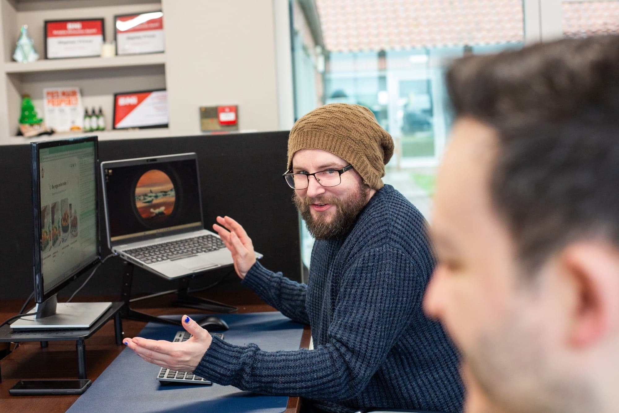A man in a woolly hat talking animatedly at his desk