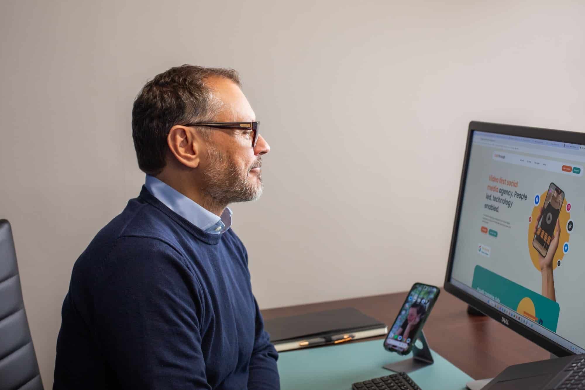 A man working at a desk, with a website on his monitor and a phone beside him