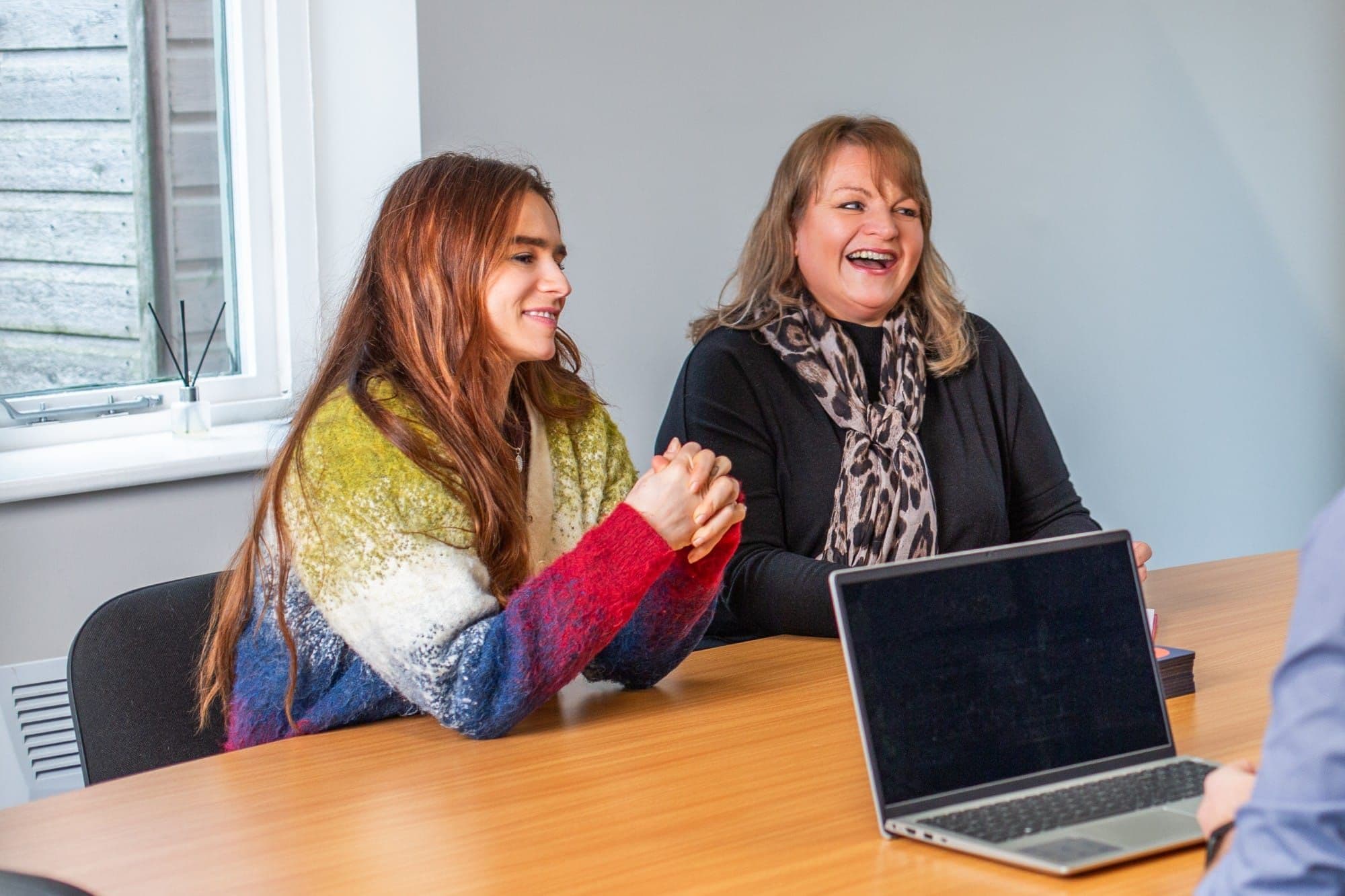 Two women laughing together at a meeting table with a laptop