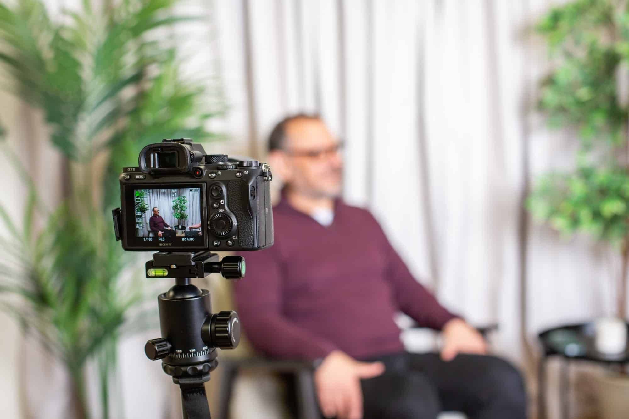 A camera on a tripod filming a man seated in the studio